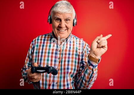 Homme de gamer hante senior jouant au jeu vidéo à l'aide de la manette et du casque très heureux pointant avec la main et le doigt sur le côté Banque D'Images