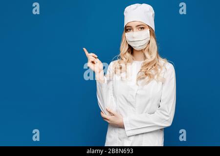 Une jeune infirmière dans un manteau blanc et un masque médical pose sur un fond bleu et pointe un doigt sur le côté gauche, isolé avec un espace de copie. Jeune femme Banque D'Images