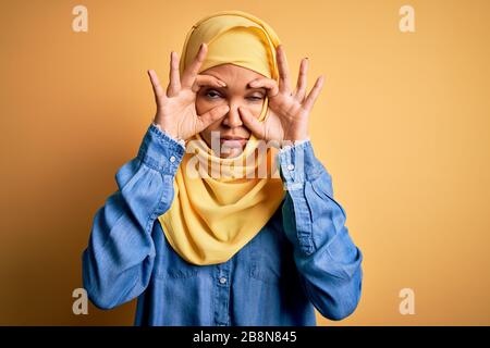 Jeune femme belle avec des cheveux bouclés portant le hijab traditionnel arabe sur fond jaune essayant d'ouvrir les yeux avec les doigts, endormie et fatigué pour le tremblement Banque D'Images