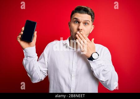 Jeune homme d'affaires aux yeux bleus montrant écran de smartphone sur fond rouge couvrir la bouche avec la main choquée de honte pour l'erreur, l'expression de fe Banque D'Images