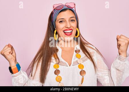 Jeune femme hippie hispanique portant le style boho de la mode et des lunettes de soleil sur fond rose célébrant surprise et étonné de succès avec des rais d'armes Banque D'Images