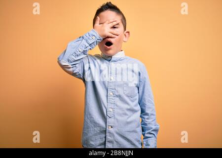 Jeune garçon enfant portant une élégante chemise debout sur un fond jaune isolé peeking dans le visage et les yeux de revêtement de choc avec la main, regardant à travers Banque D'Images