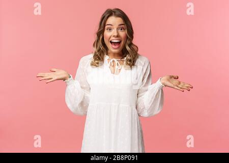 Surprise et heureuse jeune fille blonde réaliser qu'elle a gagné, recevoir vraiment de bonnes nouvelles, se répandre les mains sur le côté souriant et regarder étonné de l'appareil photo, debout rose Banque D'Images