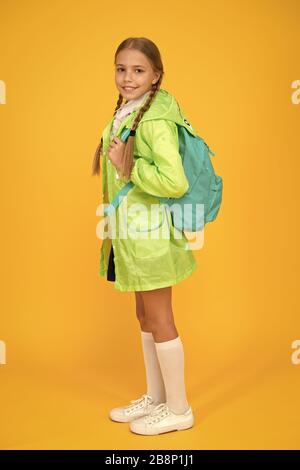 Sur votre dos et prêt à aller à l'école. Petite fille de l'école porter imperméable mignon sur fond jaune. Adorable gamin avec sac à dos pour l'école de jour de pluie. Petit Enfant retour à l'école en automne. Banque D'Images