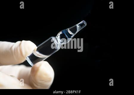 Ampoule en verre de médicament tenue par la main gantée sur fond noir. Banque D'Images