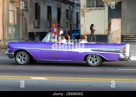 Voiture violette classique avec les touristes passant devant des constructions en mauvais état à Malecon Promenade à la Havane, Cuba. Véhicule vintage aux couleurs éclatantes Banque D'Images