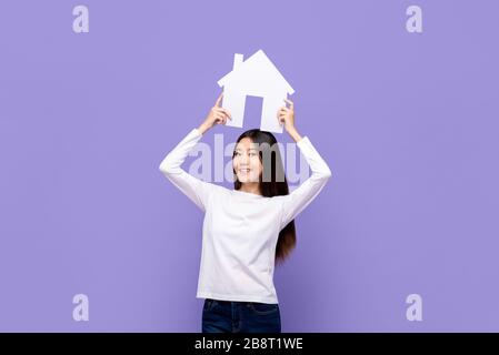 Belle femme asiatique souriante tenant le symbole de la maison au-dessus isolé sur fond mauve Banque D'Images