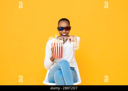 Une heureuse femme africaine américaine avec des lunettes en relief et du pop-corn riant tout en regardant un film sur fond de studio jaune Banque D'Images