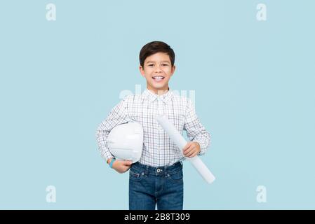 Jeune garçon asiatique de race mélangée souriant aspirant à être ingénieur tenant photocalque et casque isolé sur fond bleu clair de studio Banque D'Images