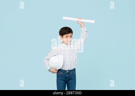 Joli jeune garçon asiatique souriant avec casque aspirant à être futur ingénieur montrant photocalque isolé sur fond bleu clair de studio Banque D'Images