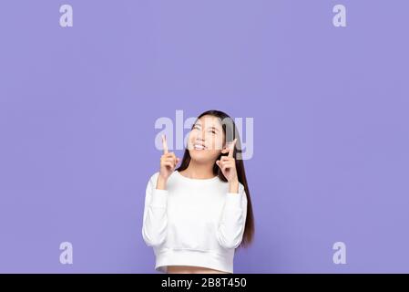 Portait d'une jeune femme asiatique souriante mignonne pointant vers le haut et regardant un espace vide au-dessus dans un fond de studio isolé pourpre Banque D'Images