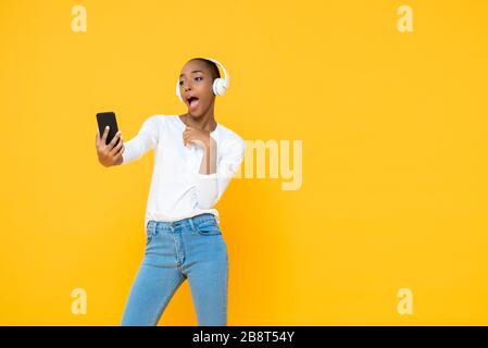 Une femme afro-américaine confiante portant un casque à l'écoute de la musique du téléphone portable dans un fond de studio isolé en jaune Banque D'Images