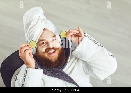 Homme à la graisse drôle barbu avec un masque cosmétique sur son visage dans peignoir serviette sur sa tête sur son visage reposant sur un fond gris Banque D'Images