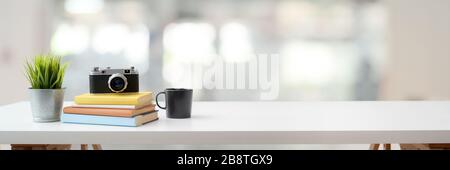 Prise de vue rognée d'un espace de travail confortable avec appareil photo, livres, tasse à café, cafetière et espace de copie sur une table blanche avec arrière-plan flou Banque D'Images