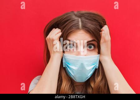Jeune femme effrayée dans un masque médical debout sur fond rouge. Banque D'Images