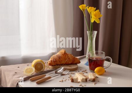 Dans la chambre sur la table près de la fenêtre avec des rideaux blancs et bruns, le petit déjeuner est servi avec une tasse de thé, des croissants, des citrons et des jonquilles jaunes i Banque D'Images