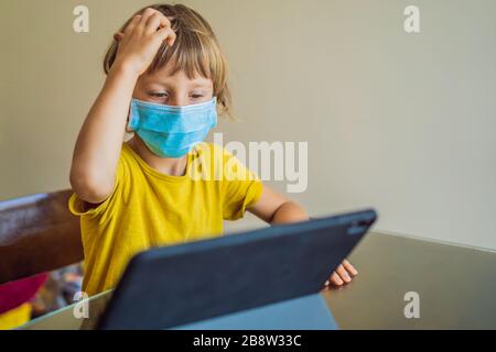 Garçon étudiant en ligne à la maison à l'aide d'une tablette. Maman l'aide à apprendre. Maman et fils dans des masques médicaux pour protéger contre le coronovirus. Étudier pendant Banque D'Images