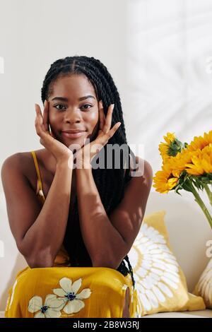 Jeune femme positive avec des readlocks assis sur un canapé dans le salon, touchant le visage et regardant l'appareil photo Banque D'Images