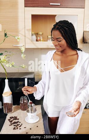 Femme souriante en vêtements de nuit mettant une cuillère de café moulu pour préparer une boisson de style vietnamien le matin Banque D'Images
