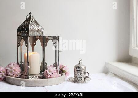 Tasse de thé décorative en argent, cerisiers roses en fleurs et lanterne marocaine en lin sur la table près de la fenêtre. Dîner iftar. Ramadan Kareem gr Banque D'Images