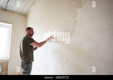 Le travailleur de l'homme fait la réparation dans l'appartement en mettant la putty sur les murs. Jeune homme avec une barbe dans un t-shirt et un short enduit de peinture travaille jour. Maison de construction. Banque D'Images