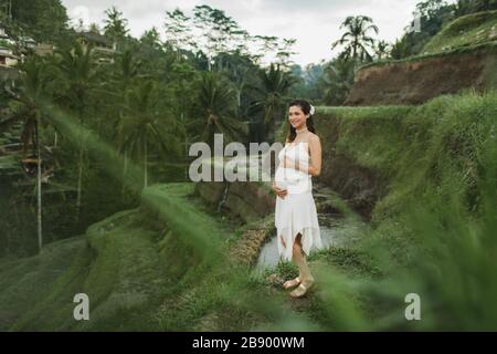 Jeune femme enceinte en robe blanche avec vue sur les rizières en terrasses de Bali dans la lumière du soleil du matin. Harmonie avec la nature. Concept de la grossesse. Banque D'Images