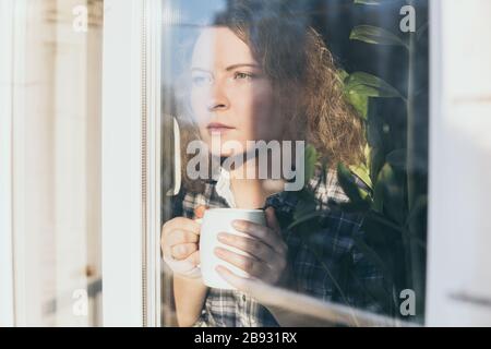 Jeune femme blonde regardant hors de la fenêtre avec une expression concernée sur son visage. Banque D'Images