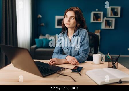 Jeune fille assise à la table avec ordinateur portable et regardant loin Banque D'Images