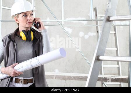femme constructeur de travailleurs de la construction parler de téléphone mobile et tenant bluprint, porter casque et casque de protection auditive dans le bâtiment à l'intérieur Banque D'Images