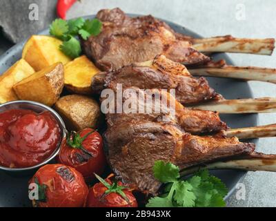 Côtelettes d'agneau rôties sur une planche en bois avec pommes de terre à sauce rouge et tomates Banque D'Images