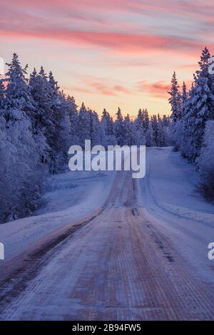 Paysage d'hiver d'une route longieuse menant à travers une forêt en Laponie au coucher du soleil, Finlande Banque D'Images