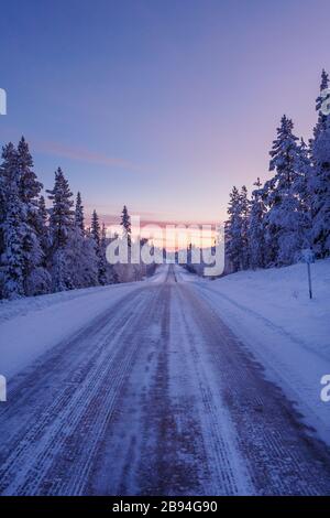 Paysage d'hiver d'une route longieuse menant à travers une forêt en Laponie au coucher du soleil, Finlande Banque D'Images