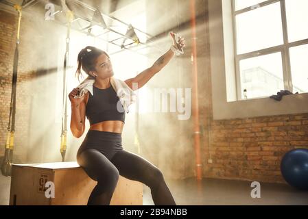 Prendre une photo après l'entraînement. Bonne femme sportive dans un casque portant une serviette sur ses épaules et faisant du selfie tout en étant assise à la salle de sport. Technologies numériques. Concept sportif. Concept de photo Banque D'Images