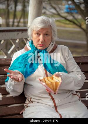 Une femme âgée, triste, habillée avec élégance, est assise sur un banc de parc avec un sac à main vide dans ses mains Banque D'Images