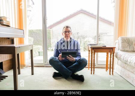 50 quelque chose de mâle dans l'auto-isolation du coronavirus à la maison, assis devant la fenêtre dans le salon, en regardant tablette ordinateur pad Banque D'Images