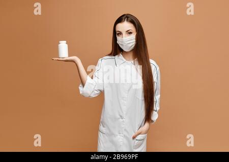 Jeune femme en vêtements médicaux et masque de protection posant avec pot de pilules, isolé sur fond beige. Le médecin féminin garde dans les pilules pour la main. Virus Banque D'Images