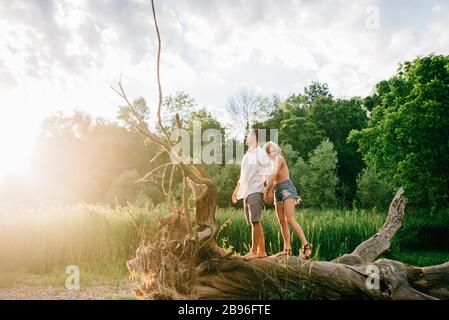Jeune couple debout sur un arbre tombé sur une rive, tenant les mains et souriant. Banque D'Images