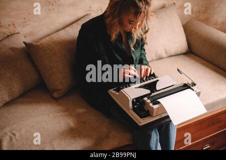 Jeune écrivain féminin utilisant la machine à écrire tout en étant assis sur un canapé à la maison Banque D'Images