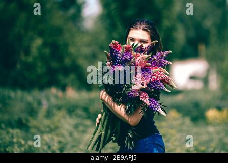 Une jeune femme marche dans le pré parmi l'herbe verte et les lupins pourpre avec un bouquet de lupins à la main Banque D'Images