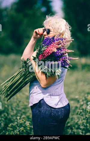 Blonde femme mûre tenant un bouquet de lupin sur le fond de la nature. Le concept de beauté et de corps positif. Coucher de soleil Banque D'Images