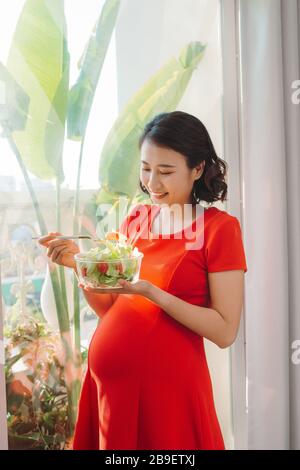 une jeune femme enceinte heureuse mangeant une salade de légumes et souriant près de la fenêtre à la maison Banque D'Images