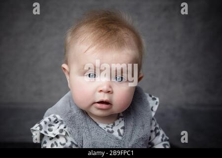 portrait facial d'une petite fille de 7 mois face à la caméra et sur un fond gris portant une tenue grise. Banque D'Images