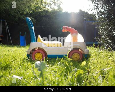 Le plastique de l'enfant s'assoit sur le jouet dans le jardin ensoleillé après la pluie avec une lentille torchère Banque D'Images