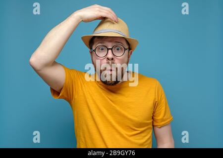 Surpris l'homme caucasien dans des verres ronds s'accroche à son chapeau, comme si saluant quelqu'un, sur un fond bleu. Banque D'Images