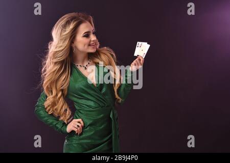 Blonde femme dans une robe verte et élégante et bijoux. Mettez la main sur la hanche, souriant, montrant deux as, posant sur fond mauve. Poker, casino. Gros plan Banque D'Images