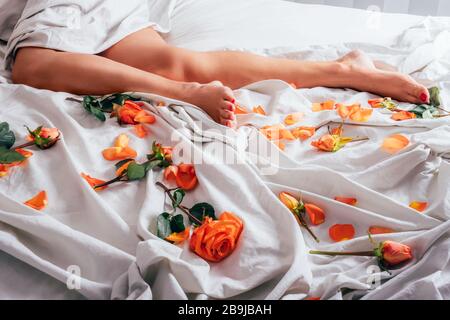 Vue courte des jambes de la femme reposant sur une literie blanche et douce avec des roses et pétales rouges oranges. Banque D'Images