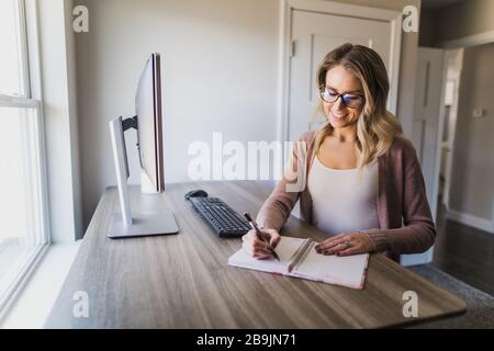 Jeune femme avec des lunettes écrivant dans son carnet dans son bureau à la maison Banque D'Images