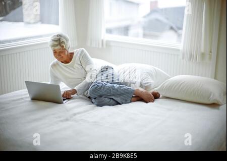 Une femme mûre dans son pyjama sourit légèrement quand elle se trouve sur un lit penchant sur son coude et regarde vers le bas un ordinateur portable. Banque D'Images