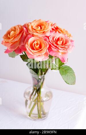 Vase en verre avec beau bouquet de fleurs de roses naturelles fraîches sur une table recouverte de tissu blanc contre un mur gris clair. Banque D'Images
