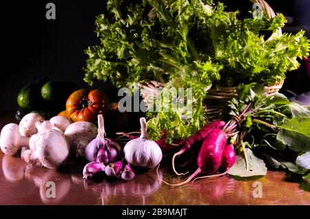 Image d'une vie encore de légumes avec un fond sombre dans lequel nous voyons l'ail violet, les champignons, les radis, les lettuces, les tomates, les concombres sur un verre Banque D'Images